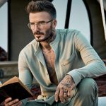 david beckham in an open olive-green shirt, holding a book and wearing glasses
