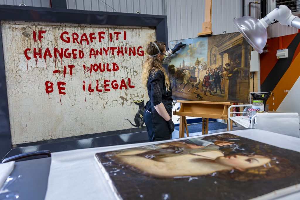 A conservator cleaning a painting on an easel with a Banksy piece behind her