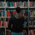 Man in black jacket with grey hoodie over blue jeans looking at a bookshelf of books in a library